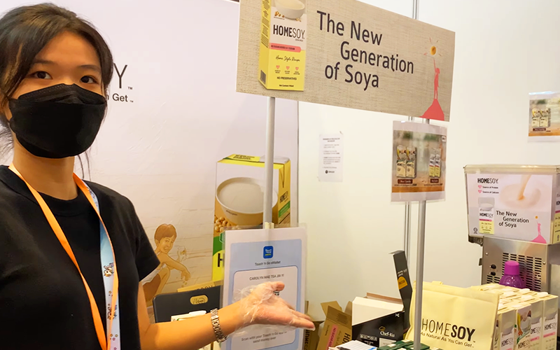 Seorang pengendali gerai ekspo makanan wanita yang memakai topeng muka kelihatan sedang menunjuk ke arah pameran produk and sampel dari jenama Home Soy seolah-olah menjemput ke sesi merasa.