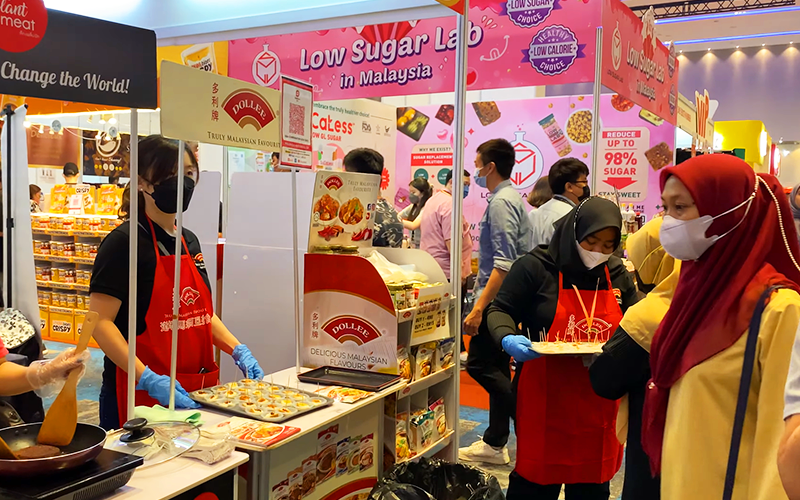 Several female staff members in face masks are stood around a food expo booth featuring a wide variety of food items that are placed on display, amidst bustling and busy foot traffic.